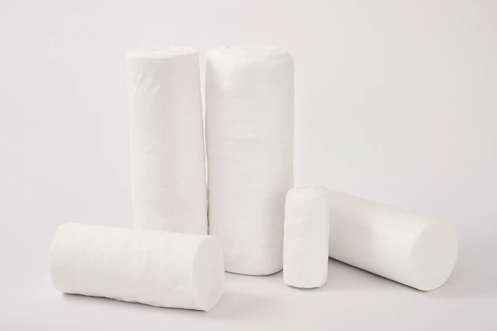 Five absorbent cotton wool rolls of different sizes, standing and lying on a grey studio background
